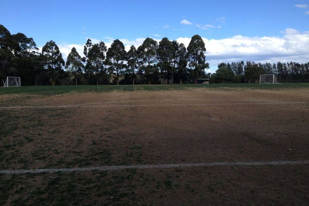 A school sports field worn to bare soil across the centre and goalmouths, with grass surviving only at the margins, a classic example of wear-dominant traffic damage